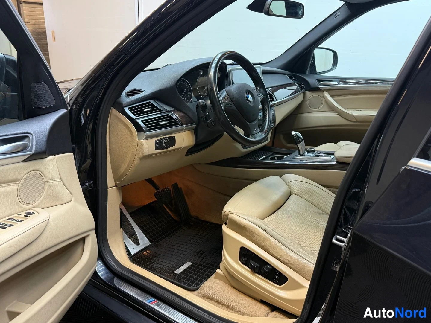 Muu: Sisätila kuljettajan ovi auki: BMW-ohjauspyörä, mittaristo, keskikonsoli ja beige nahkaistuimet; ovessa kaiutinkangas ja säätönapit.