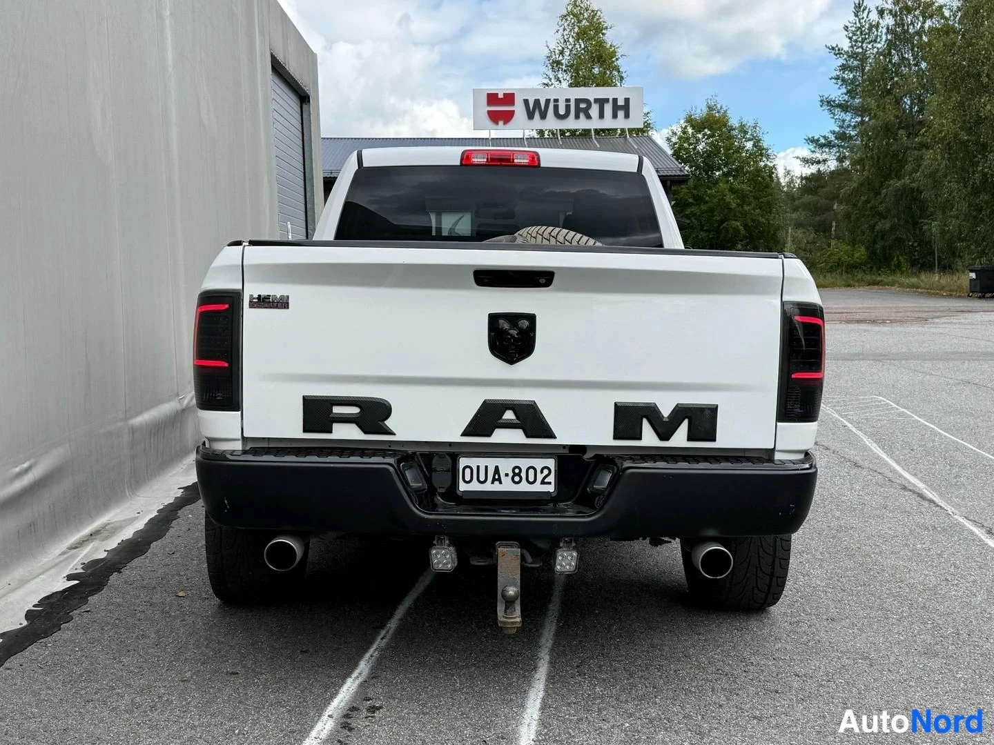 Takaa: Valkoisen lava-auton takaosa: RAM-teksti, musta merkki, rekisteritunnus, kaksi pakoputkea ja musta takapuskuri.