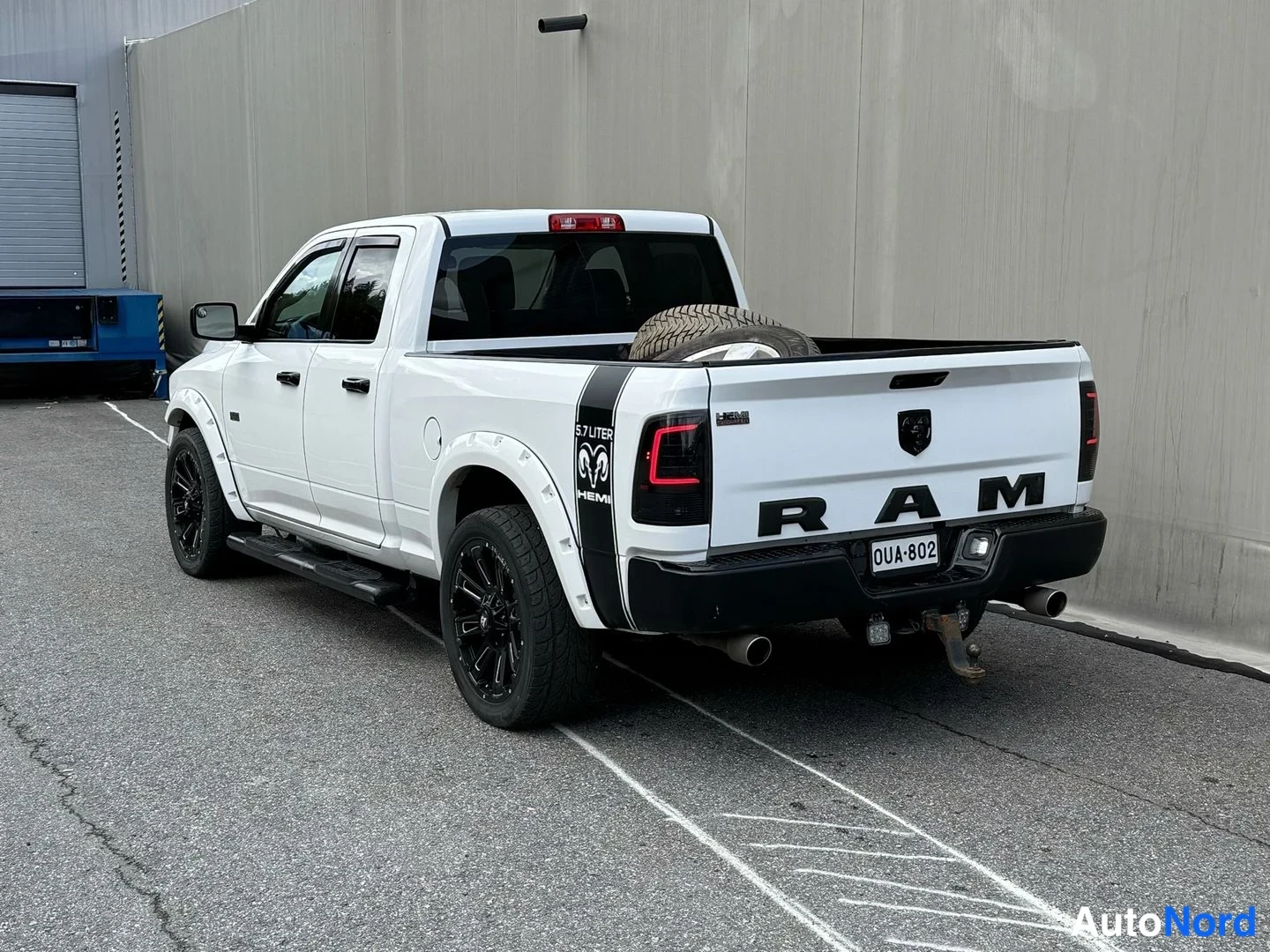 Takaviisto: Valkoinen avolava-auto (RAM 1500) takaosa, rekisterikilpi, mustat pyörät ja lava, taustalla harmaa seinä ja parkkipaikan viivat.