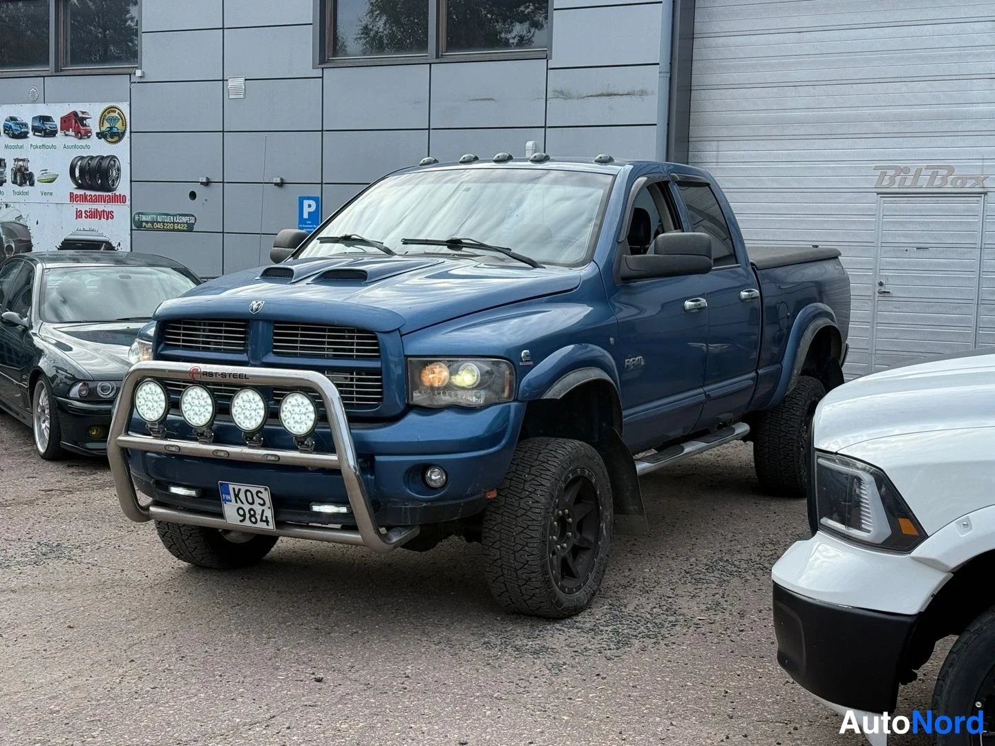 Etuviisto, nokka vasemmalle: Sininen Dodge Ram -neloautopetaksi etuviistossa, keulakaari lisävaloilla, korotettuna mustilla maastorenkailla.