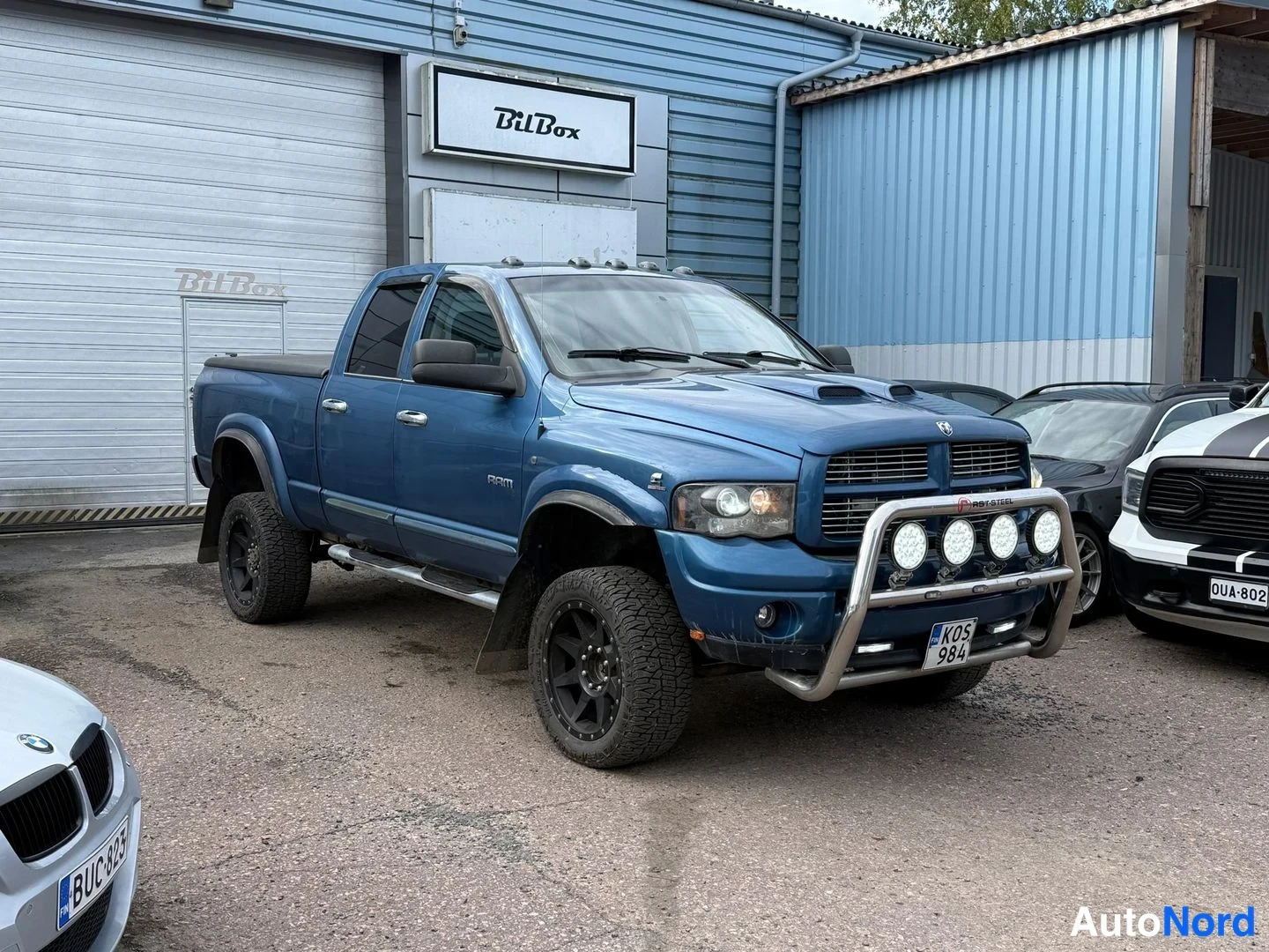 Etuviisto, nokka oikealle: Sininen pickup-auto sivulta etuviistoon oikealle; korotetut maastorenkaat, sivukaarit, taaempi lava, rekisterikilpi KOS 984.
