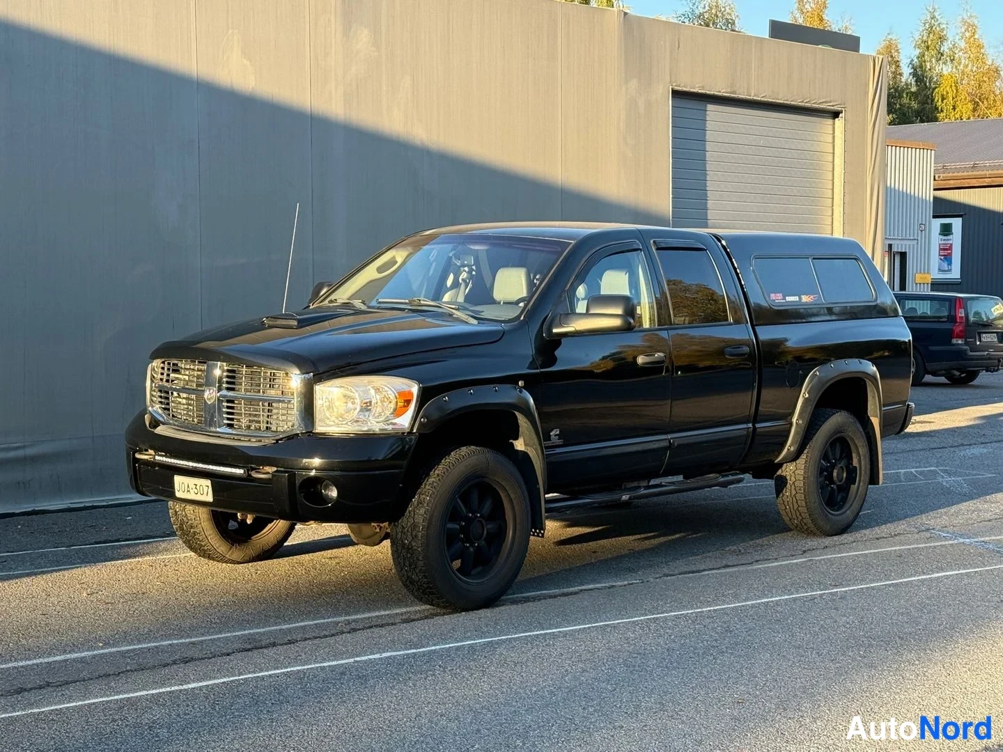Etuviisto, nokka vasemmalle: Musta Dodge Ram -nouteava avolava-auto kolmessa neljäsosassa edestä vasemmalta, mustat vanteet ja levikekaaret, lavalla tumma kova kuormatilaan asennet