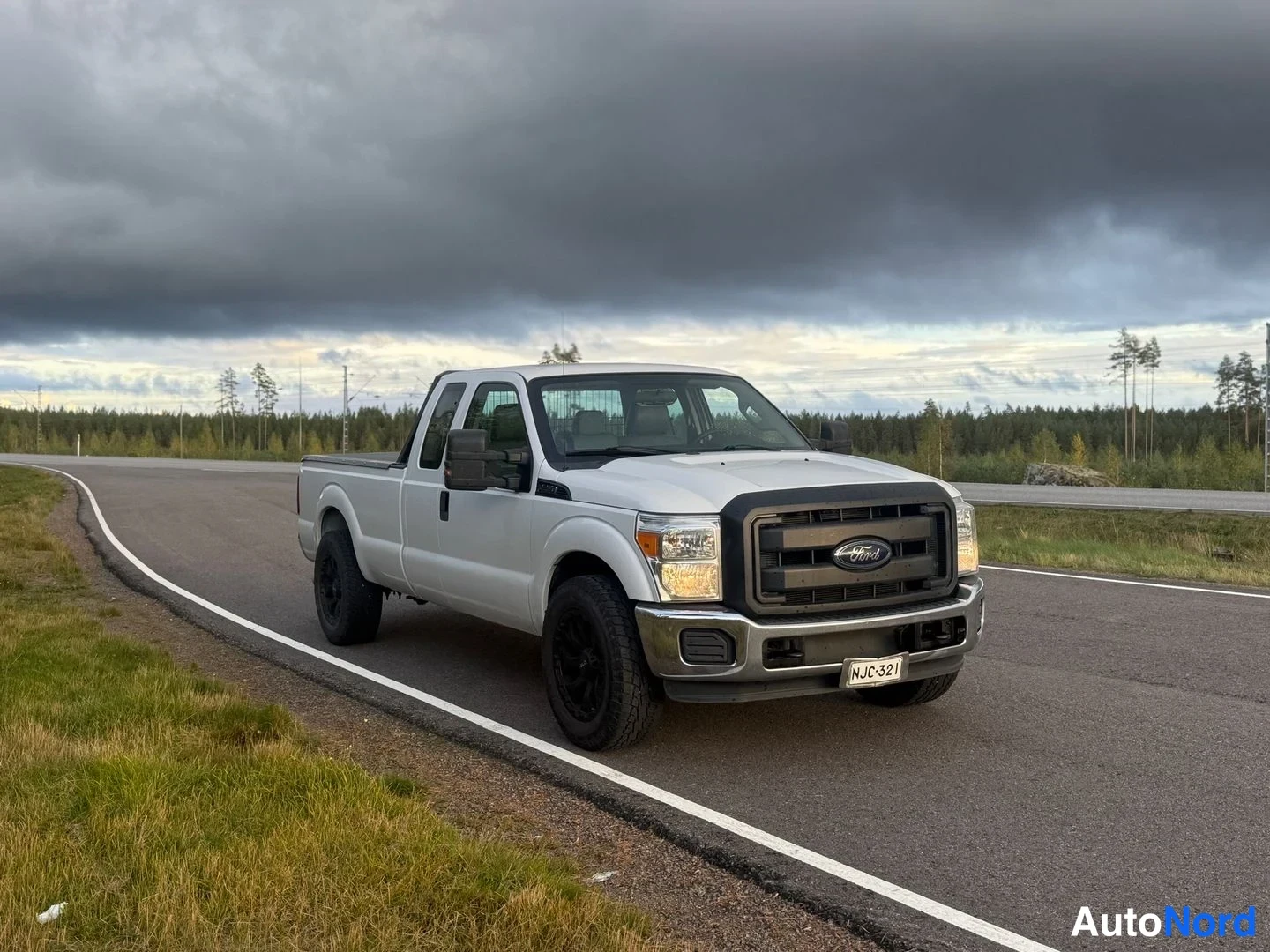 Etuviisto, nokka oikealle: Valkoinen Ford Super Duty lava-auto etuviistosta oikealta tiellä pilvisellä taivaalla.