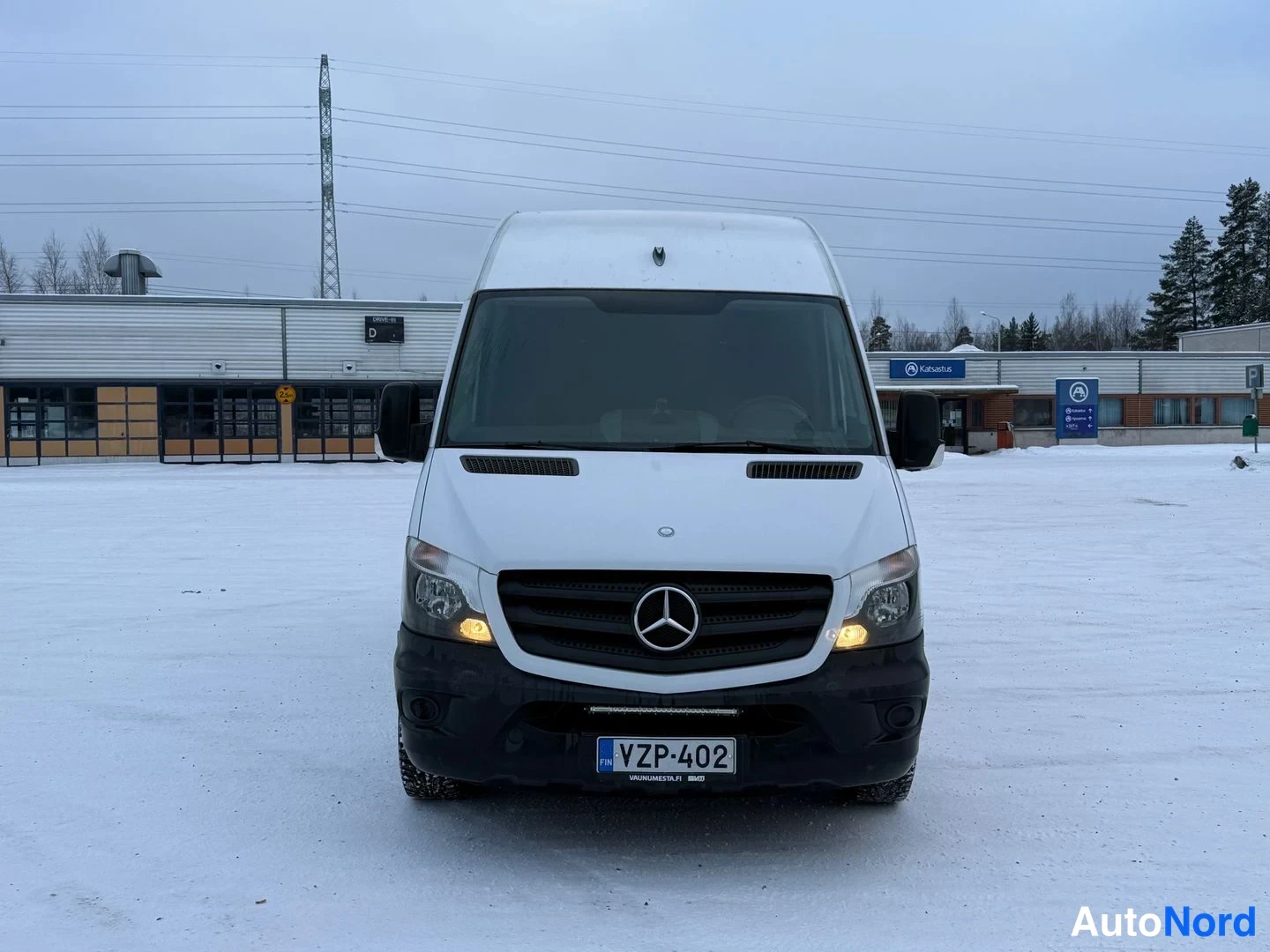 Edestä: Valkoinen Mercedes-Benz Sprinter edestäpäin otettuna lumisessa pihamaisemassa.
