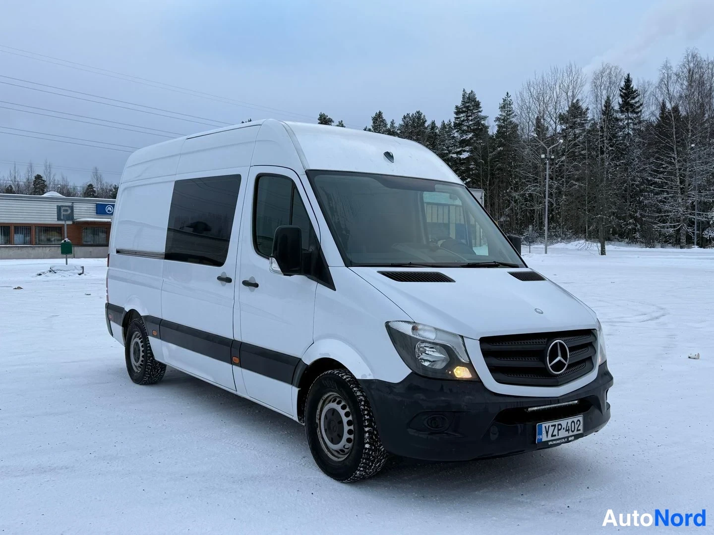 Etuviisto, nokka oikealle: Valkoinen Mercedes-Benz Sprinter etuviistossa oikealta lumisella pihalla.