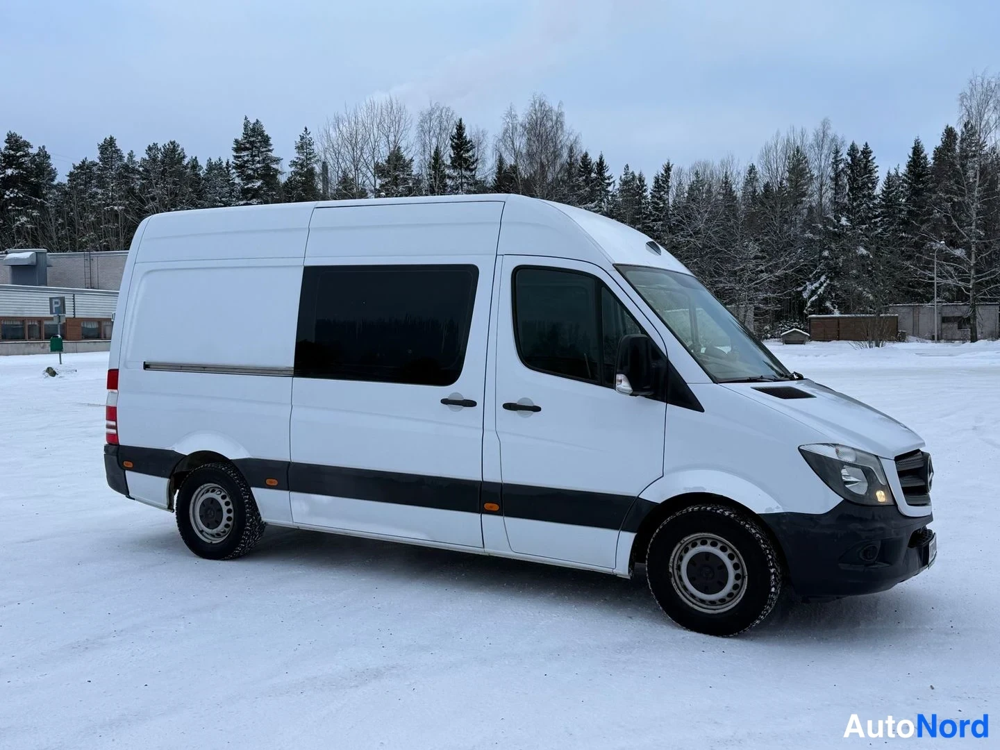 Etuviisto, nokka oikealle: Valkoinen Mercedes-Benz Sprinter -pakettiauto kolmeneljännes-kulmassa etupyörä lähellä, suojalasit ja etupuskuri näkyvissä lumisella parkkipaikalla.