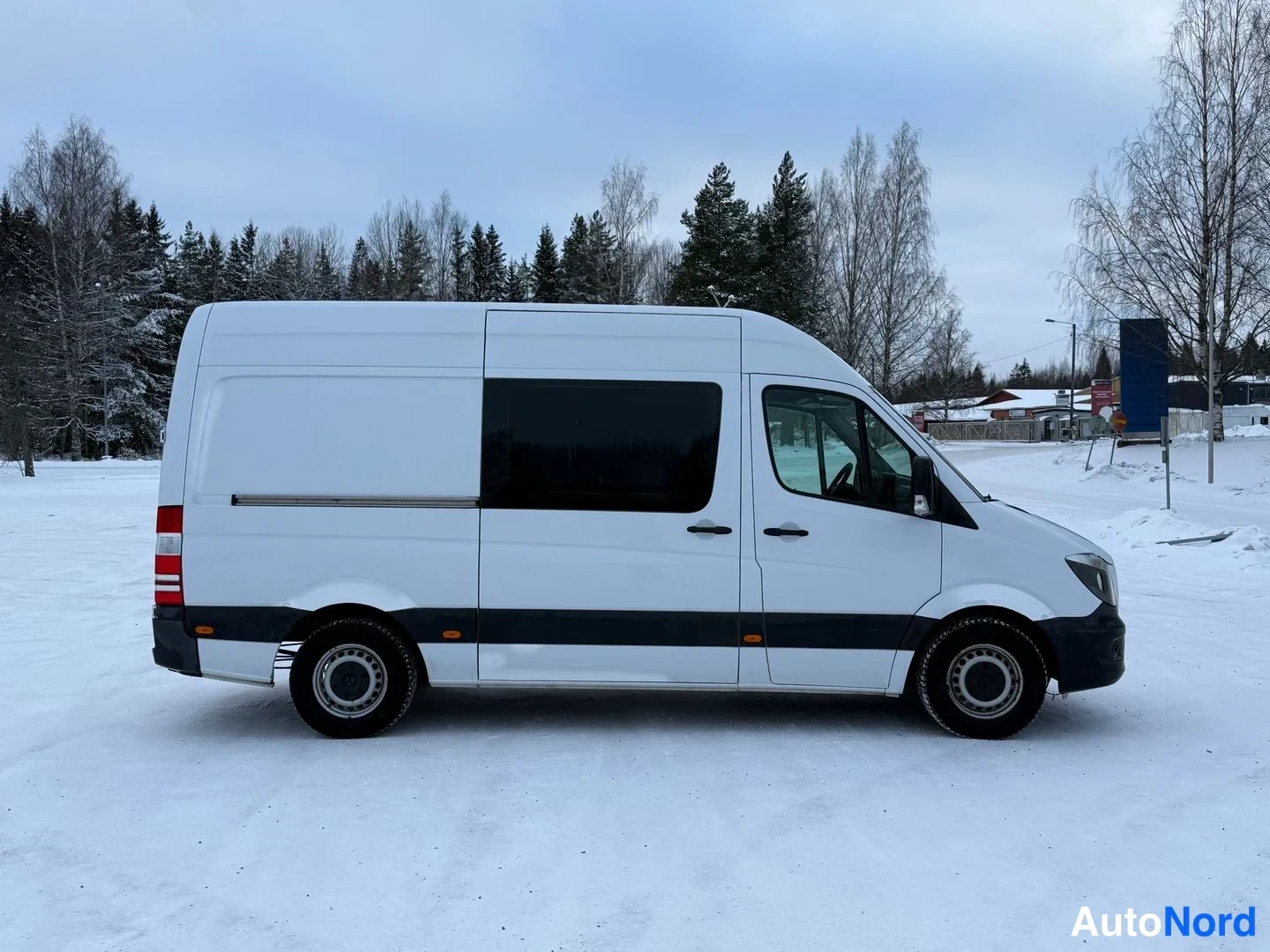 Etuviisto, nokka oikealle: Valkoinen Mercedes-Benz Sprinter -pakettiauto sivukulmassa lumen peittämällä pihalla, pitkä sivuseinä ja takavalo näkyvissä.