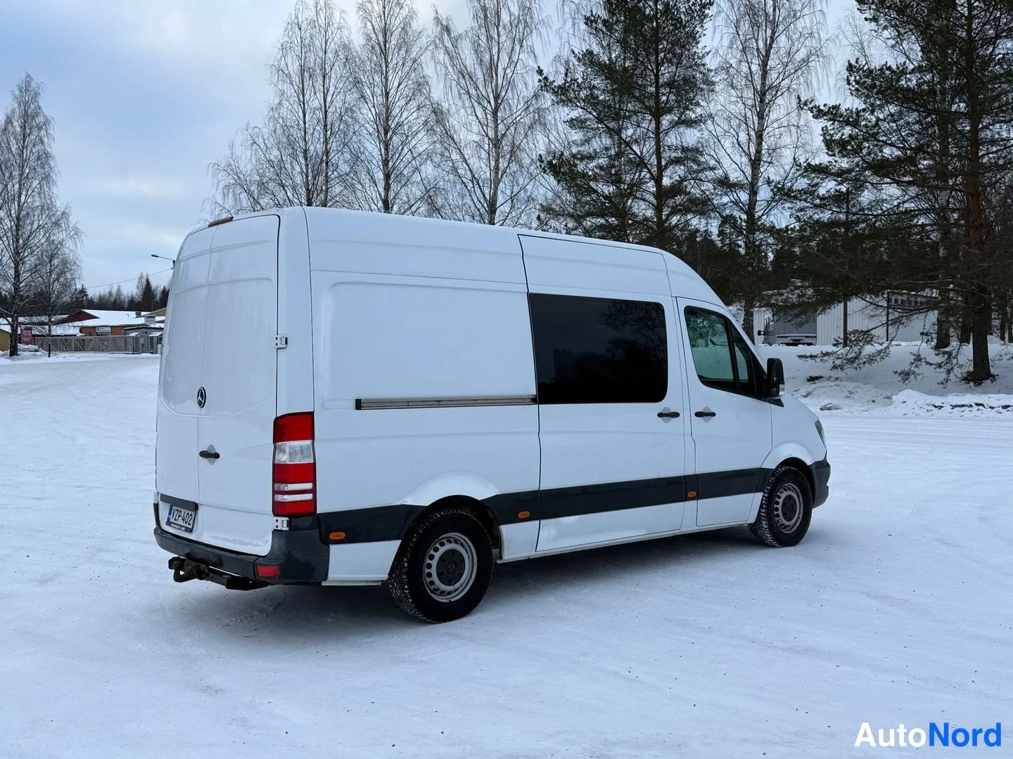 Takaviisto: Valkoinen Mercedes-Benz Sprinter -pakettiauto takakolmeneljännes-kulmassa, takaovi ja sivuoveikka näkyvät lumitillalla.