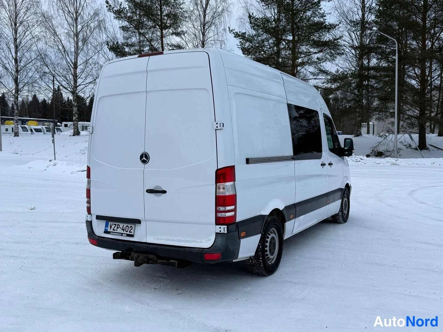 Takaviisto: Valkoisen Mercedes-Benz Sprinter -pakettiauton takaosa suoraan takaa, rekisterikilpi, vetokoukku ja Mercedes-tähti takaovessa, talvimaisemassa.