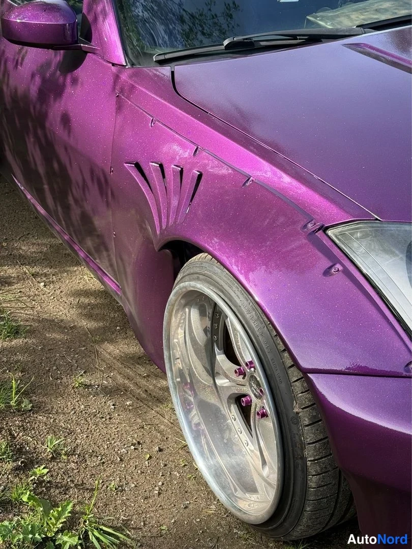Etuviisto, nokka oikealle: Lähikuva violetin auton etusivusta: pultatulla lokasuojuksella on tuuletusaukot, hopeavanne ja etupyörä, osa ajovalaisimesta oikealla.