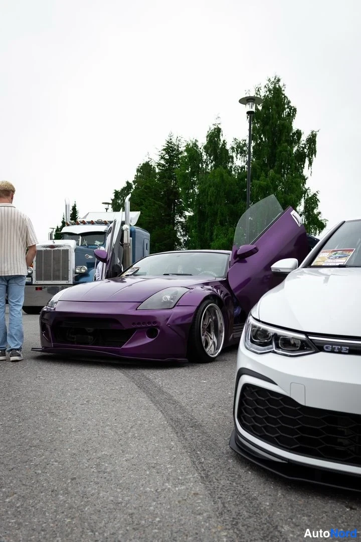 Etuviisto, nokka vasemmalle: Violetti coupé etukulmasta vasemmalla, matala etuspoileri, ovi ylhäällä; edessä osittain valkoinen auto.