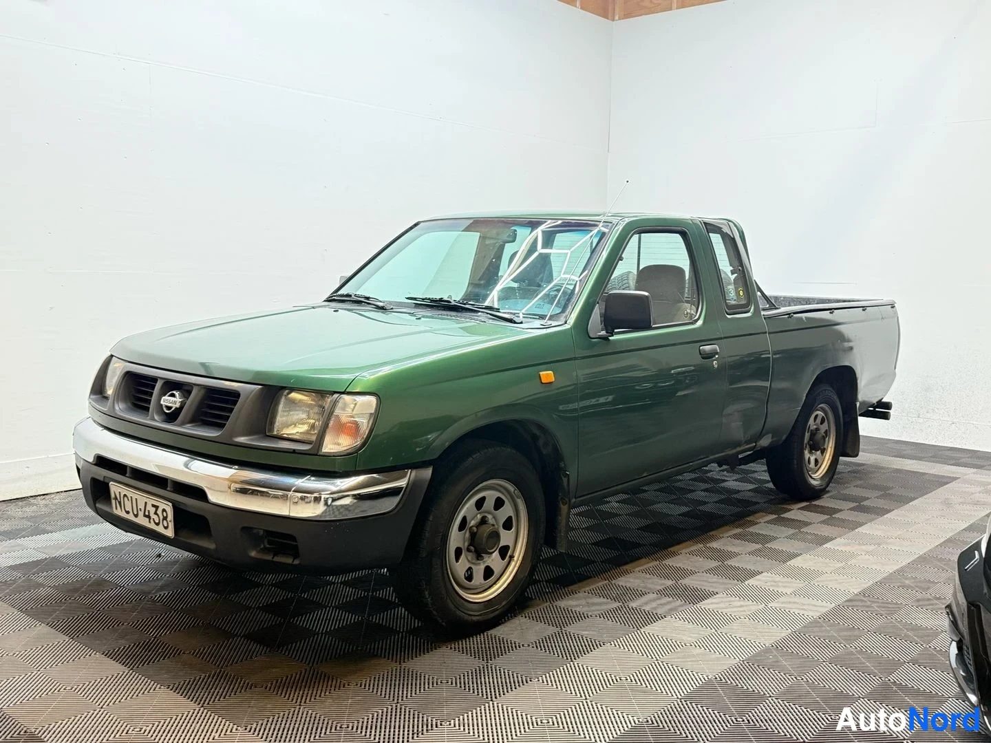 Etuviisto, nokka vasemmalle: Vihreä Nissan-m alláusauto sisätiloissa, etukulmakuva vasemmalle; rekisterikilpi ja tumma pickup-lava näkyvät.