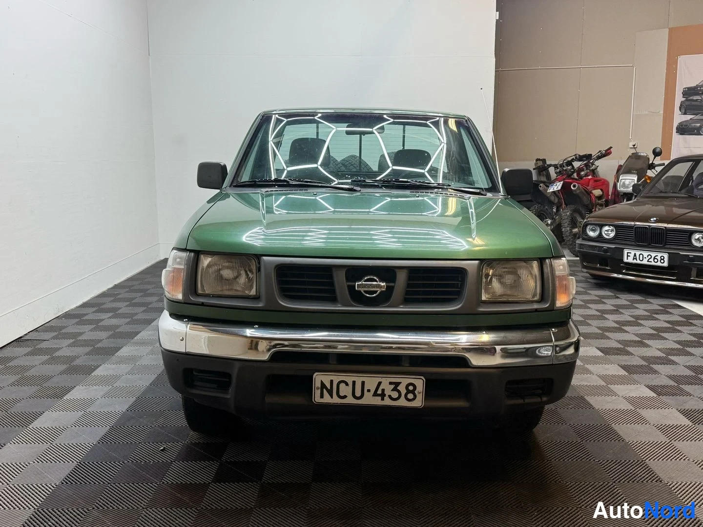 Edestä: Etukuva vihreästä Nissan-pickupista sisätilassa; kilpi, taustalla toinen auto ja moottoripyöriä.
