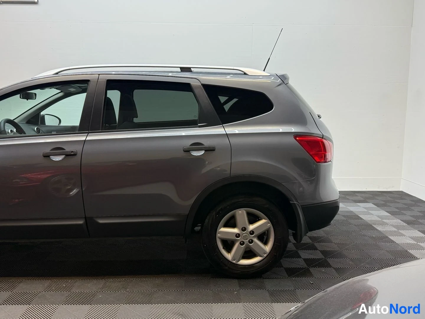 Takaviisto: Harmaa Nissan Qashqai +2 takakulmasta; perävalot, kattokaiteet ja viidennen oven takainen sivuprofiili näkyvissä sisätiloissa.