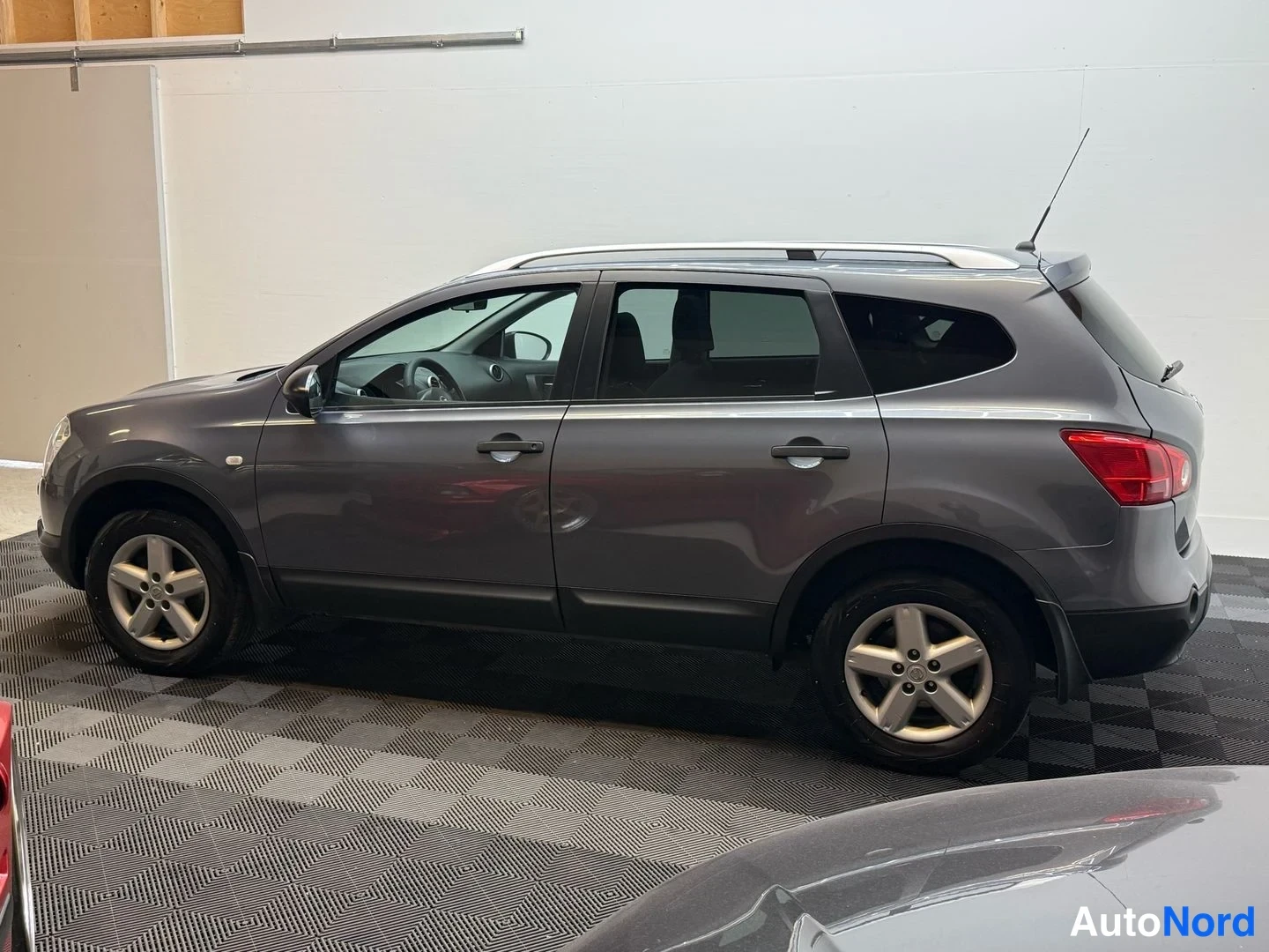 Etuviisto, nokka vasemmalle: Harmaa Nissan Qashqai +2 koko auton profiilissa etukulmasta; konepelti, ovet ja takaosa näkyvissä hallissa.