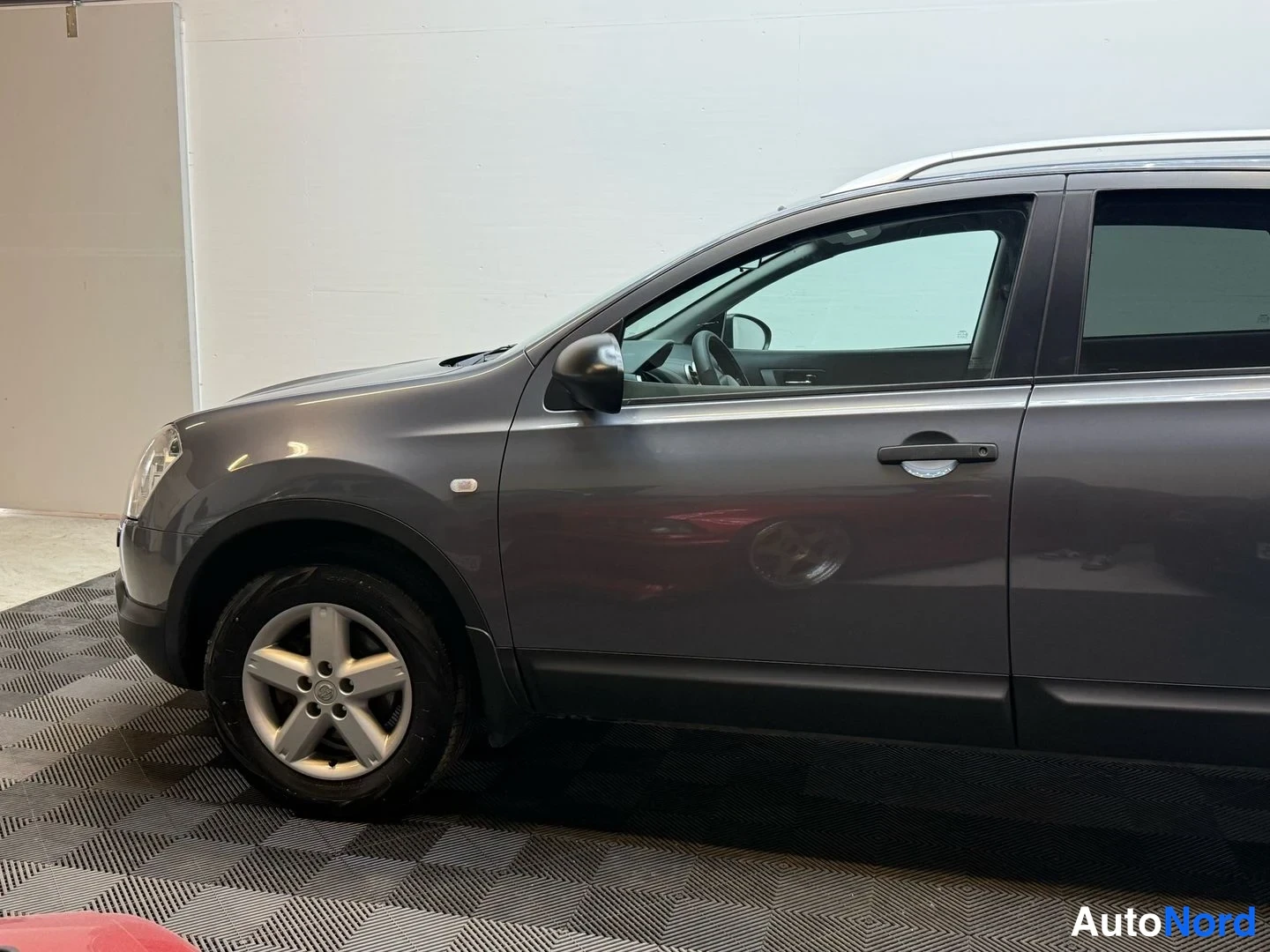 Etuviisto, nokka vasemmalle: Harmaa Nissan Qashqai +2 etukulmasta; kojelauta ja etuovet näkyvissä lavetilla.