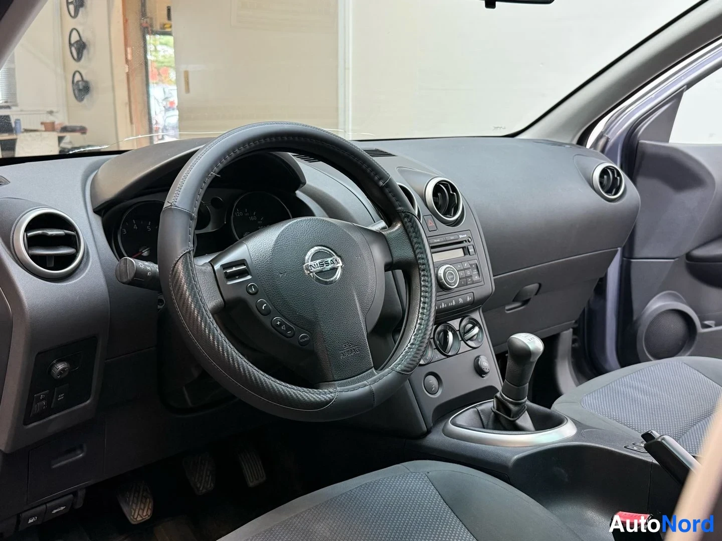 Muu: Kohtisuora kuva ohjaamosta: ratti Nissan-logolla, keskikonsoli ja manuaalivaihtehde