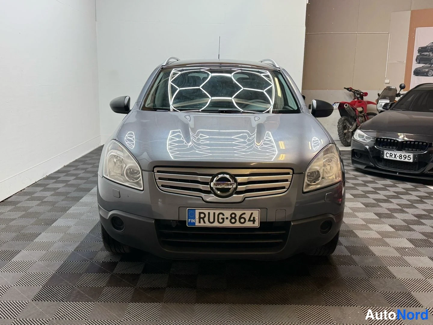 Edestä: Nissan Qashqai+2 edessäpäin, harmaa, rekisterikilpi, sisätilassa.