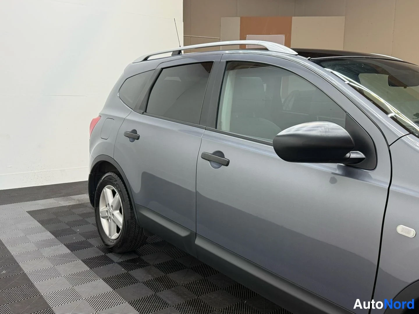 Sivulta: Nissan Qashqai+2 sivulta vasemmalta, harmaa kori, kattotelineet, mustat sivukaaret.