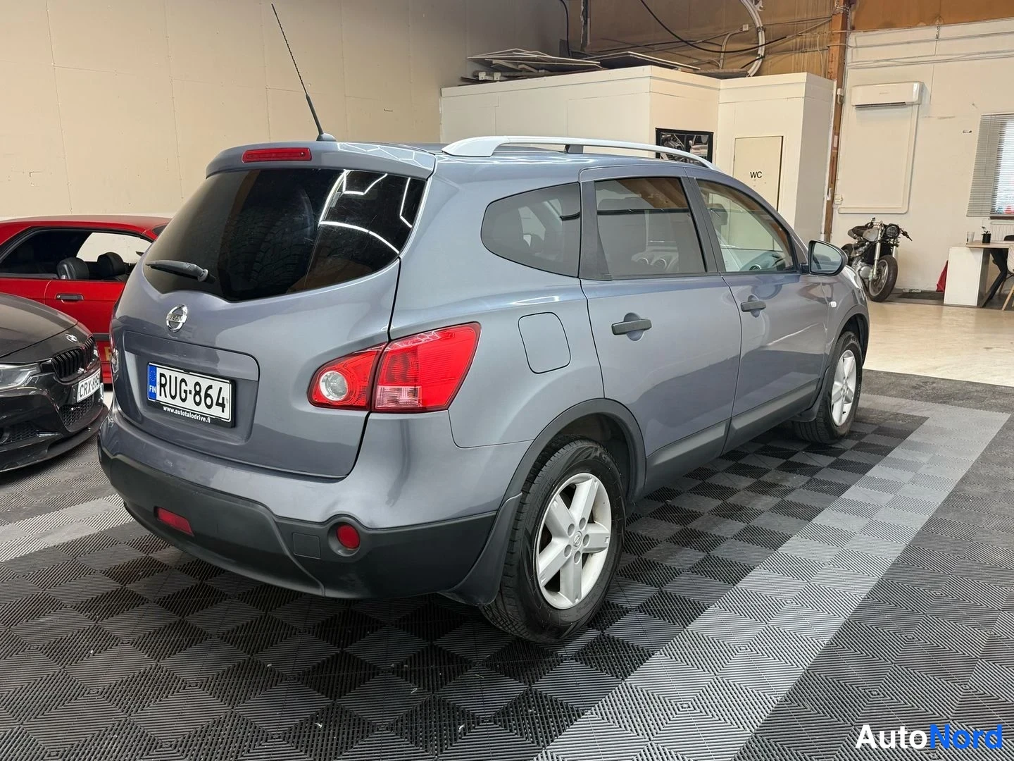 Takaviisto: Auton takaosaa ja vasenta sivua kolmessa neljäsosasta: Nissan-maasturia harmaassa hallissa, rekisterikilpi ja hopeiset kattokaaret näkyvät.