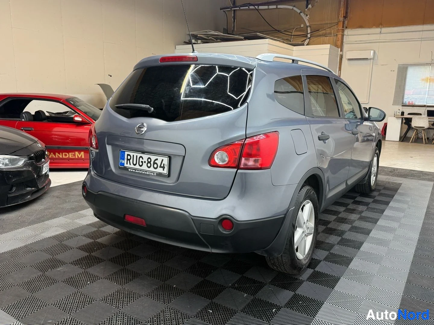 Takaviisto: Auton takaosaa ja vasenta sivua kolmessa neljäsosasta autotallissa, rekisterikilpi ja punaiset takavalot näkyvät taustalla punainen auto.