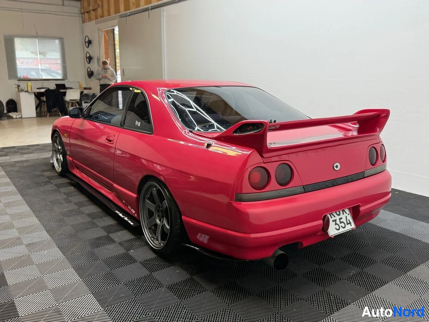 Takaviisto: Punainen Nissan Skyline takaa viistosti, takaosa ja sivuprofiili näkyvät, isot tavaratilan siivet.