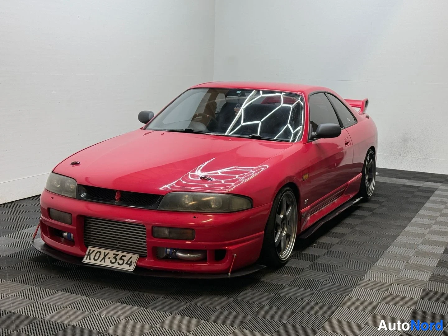 Etuviisto, nokka vasemmalle: Punainen Nissan Skyline -coupe etuviistosta vasemmalta sisätiloissa, rekisterikilpi, takaosassa suuri siipi.