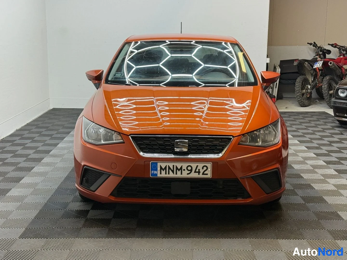 Edestä: Oranssi Seat Ibiza suoraan edestä kuvattuna sisätilassa, rekisterikilpi, taustalla moottorikelkka ja mönkijä.