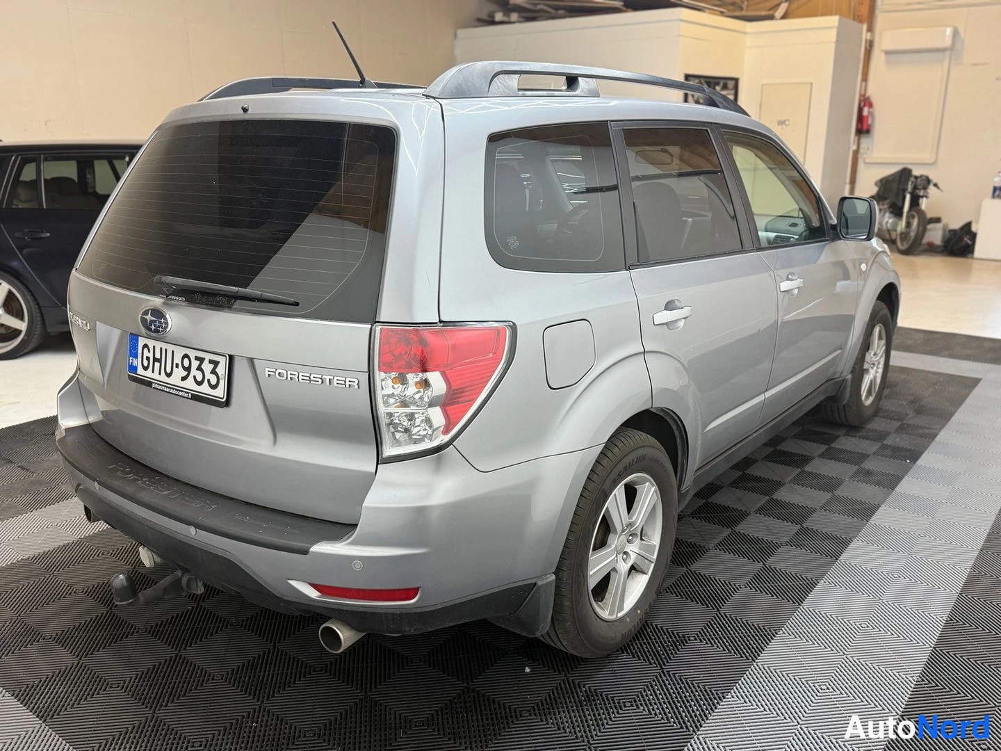 Takaviisto: Hopeanharmaa Subaru Forester takaa sivulta kuvattuna; rekisteritunnus, kattokaiteet ja takavetokoukku näkyvissä.