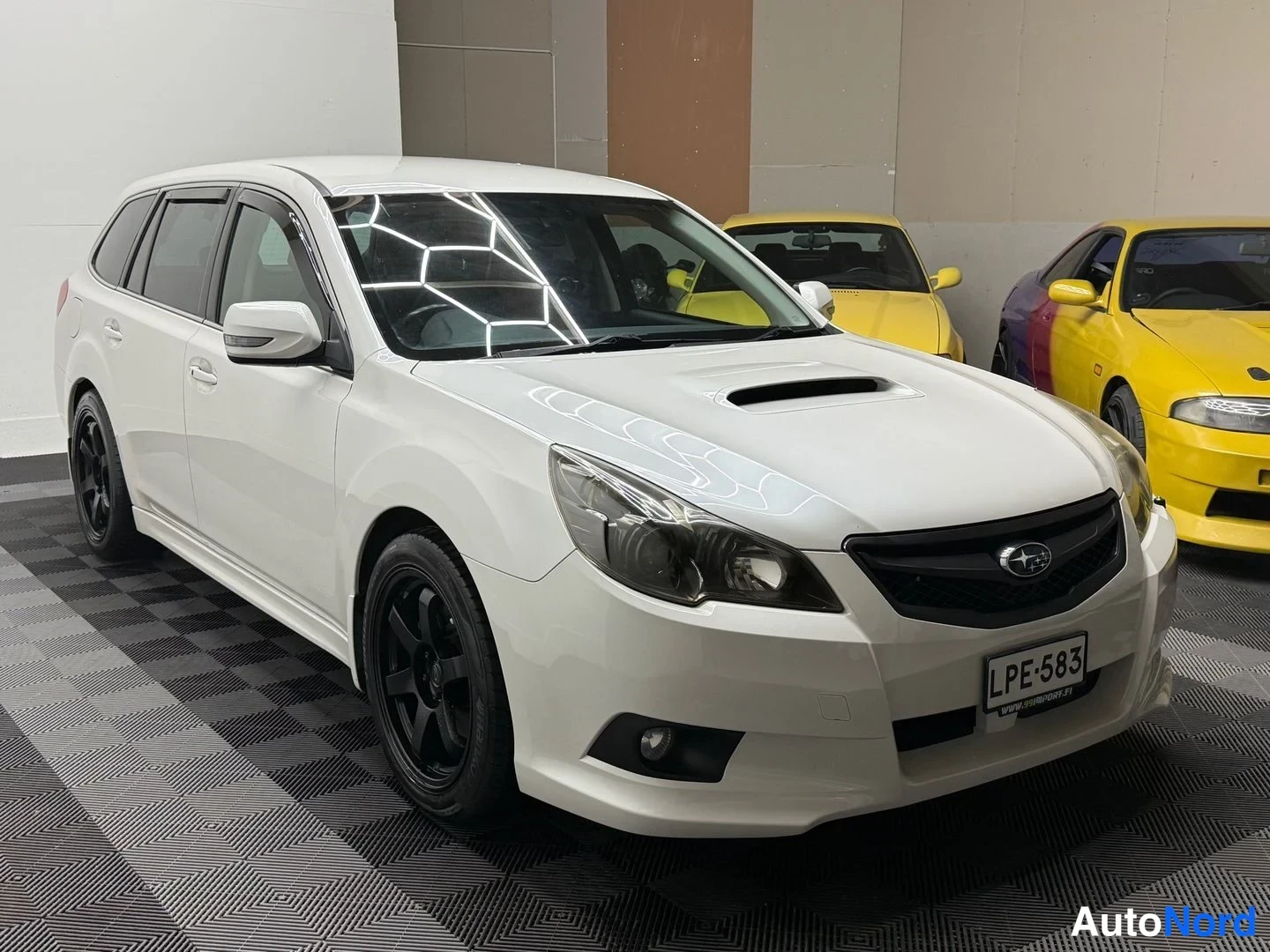 Etuviisto, nokka oikealle: Valkoinen Subaru Legacy -farmari etuviistosta oikealta, mustat vanteet ja rekisterikilpi.
