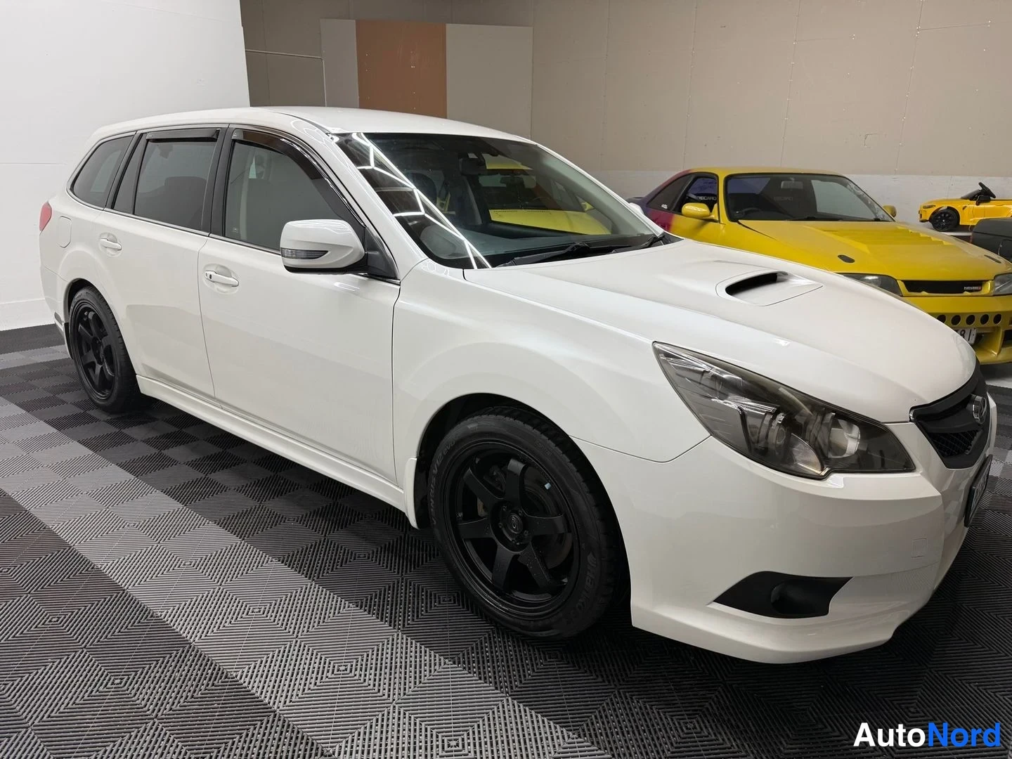 Etuviisto, nokka oikealle: Valkoinen Subaru Legacy -farmaria edestä oikealta, mustat kevytmetallivanteet ja konepelti.