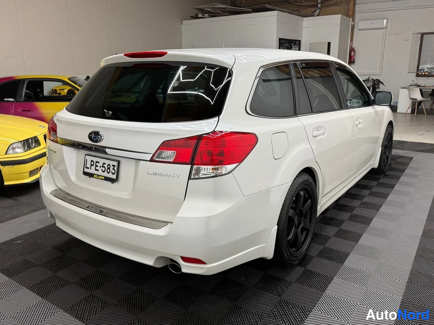 Takaviisto: Valkoisen Subaru Legacy -farmarin takapää ja oikea kylki, rekisterikilpi, mustat vanteet.