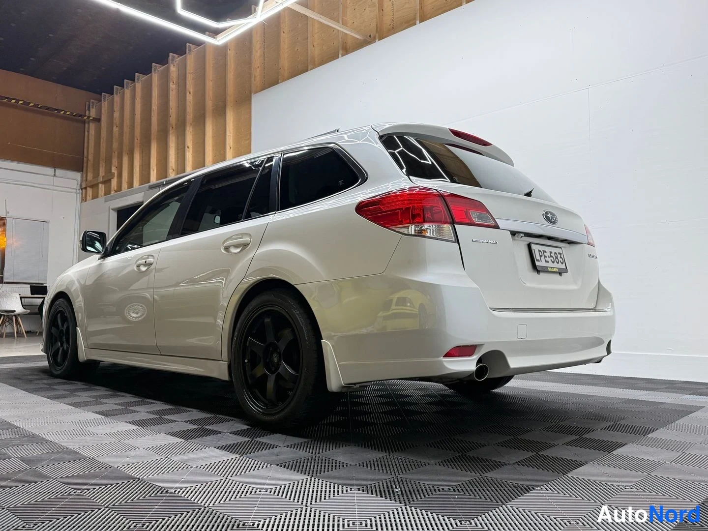 Takaviisto: Auton takakulma: valkoinen farmariauto, tummat pyörät ja rekisterikilpi sisätiloissa.