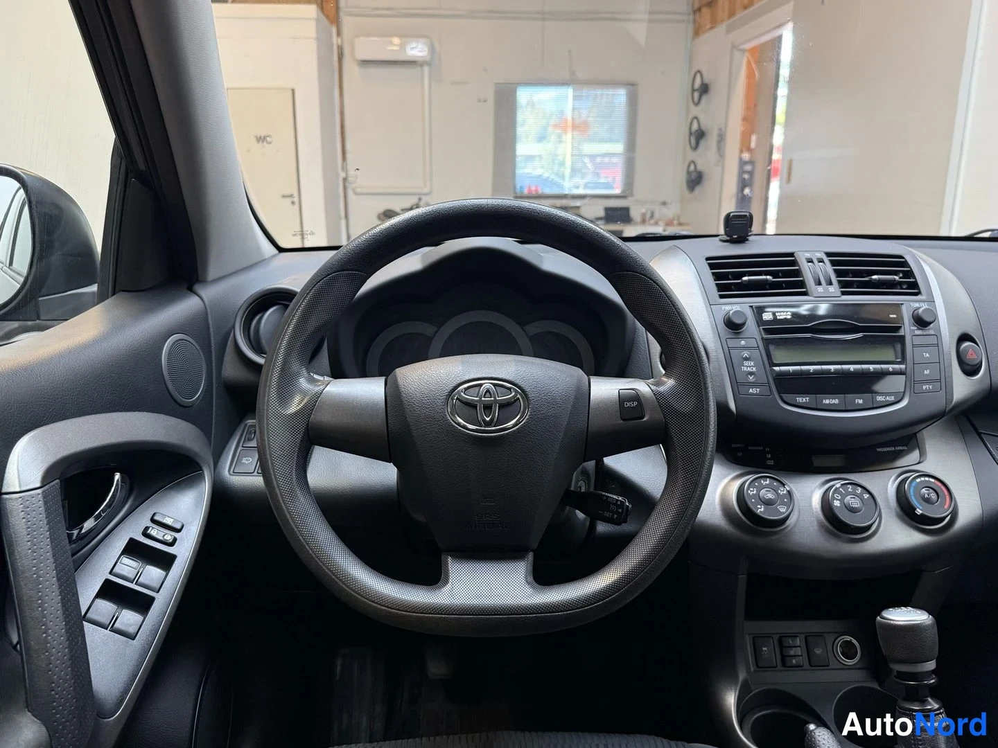 Muu: Ohjaamo suoraan edestä kuvattuna: Toyota-logo ohjauspyörässä, radioyksikkö, joka näyttää AM/FM, TEXT, AST ja DSC-AUX, sekä kolme lämmityksen säädönappia.