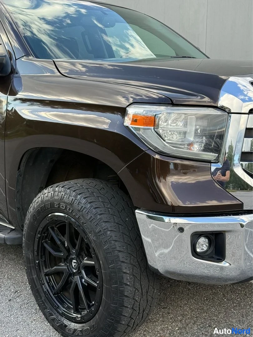 Etuviisto, nokka oikealle: Toyota Tundran etukulman lähikuva vasemman etupyörän ja ajovalon kohdalta. Musta kevytmetallivanne, TOYO OPEN COUNTRY -rengas.