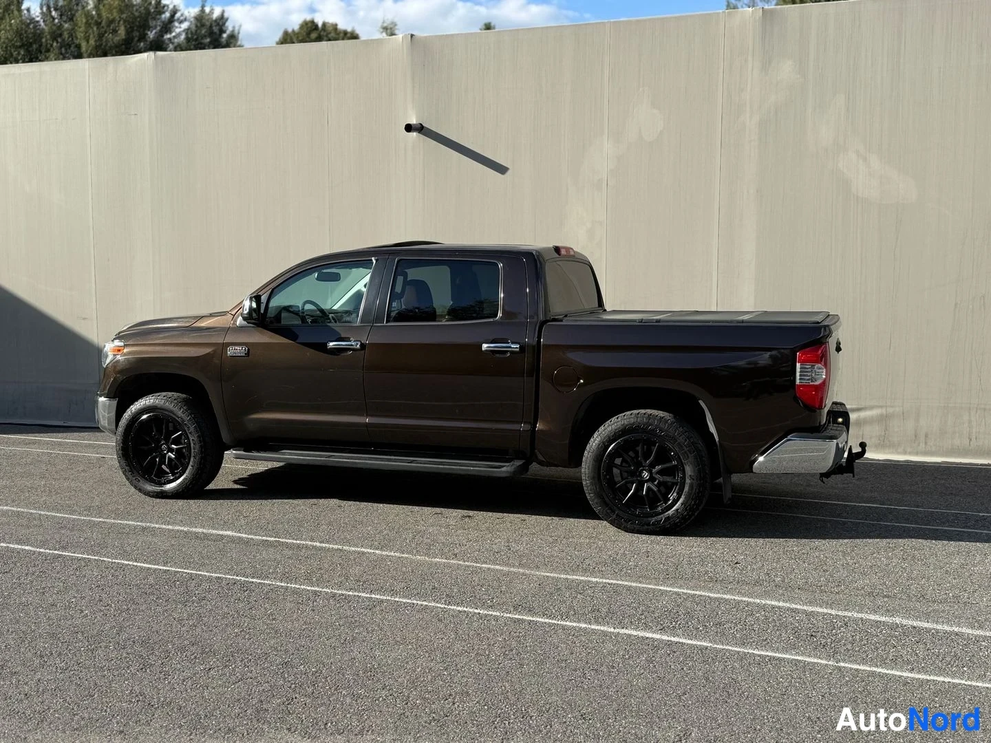 Sivulta: Toyota Tundra -lava-auto kokonaisuudessaan sivulta. Auton koriväri on ruskea, mustat kevytmetallivanteet ja kromainen takapuskuli.