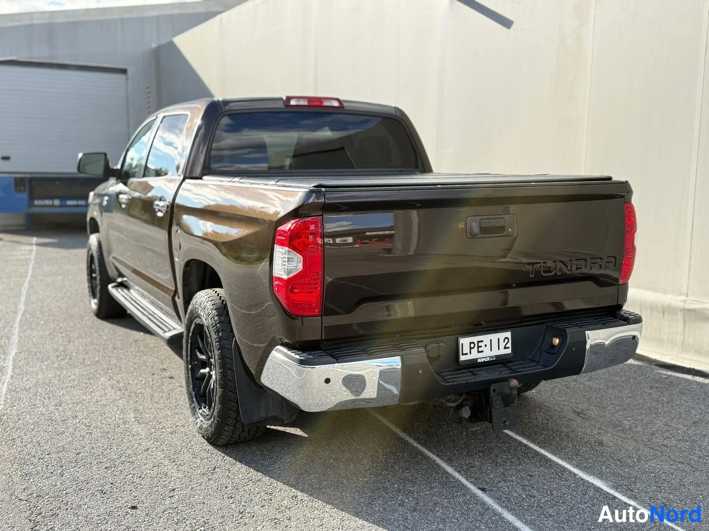 Takaviisto: Tummanruskea Toyota Tundra -nakoauton takanurkkikuva, perävaunu, TUNDRA-teksti tavaratilan kannessa, rekisteritunnus, mustat vanteet ja kromipuskuri.