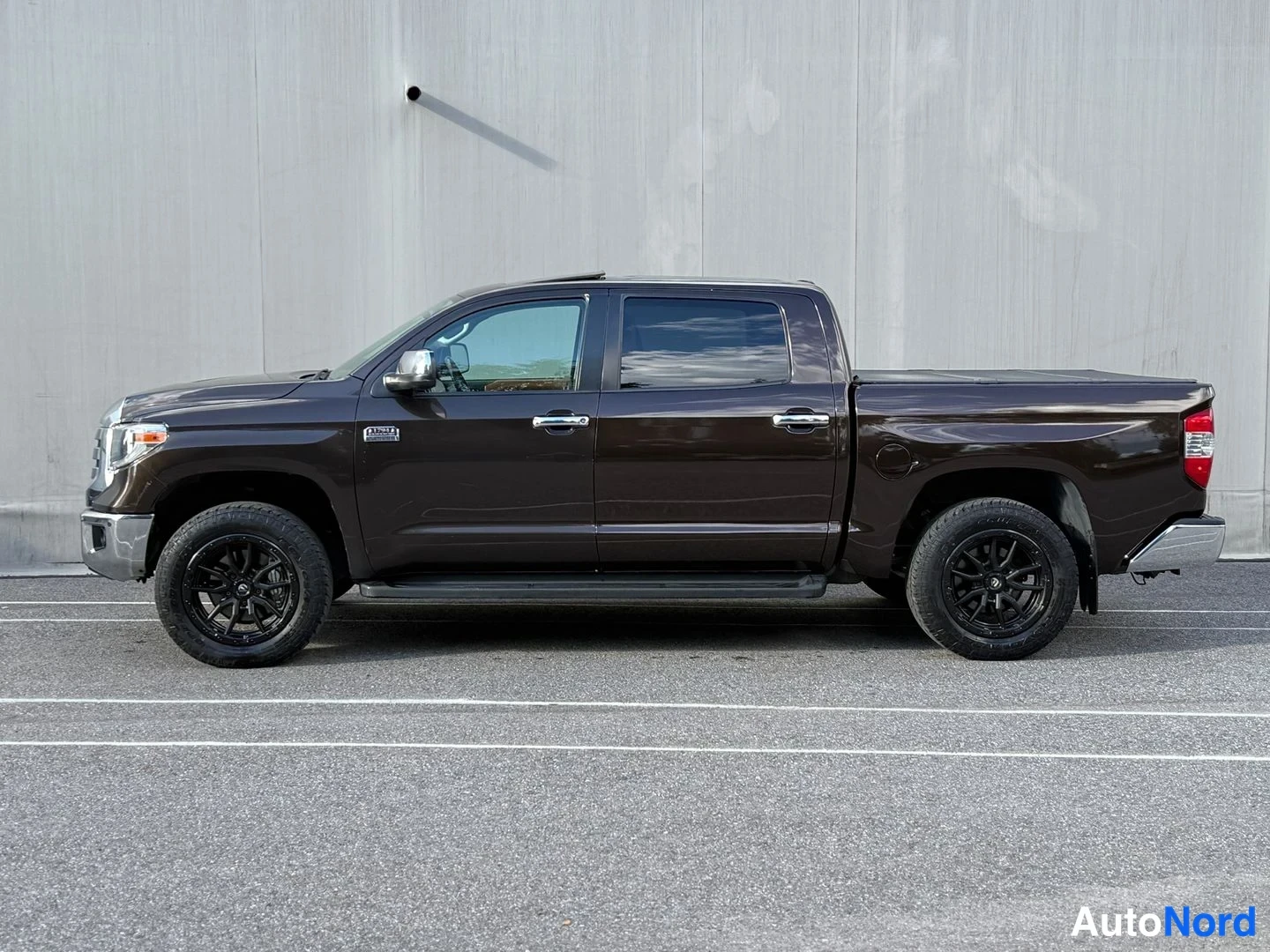Sivulta: Tummanruskean Toyota Tundra -nakoauton sivuprofiili oikealta puolen, mustat vanteet, sivukaiteet ja sähköisesti suljettu lava-kansi.
