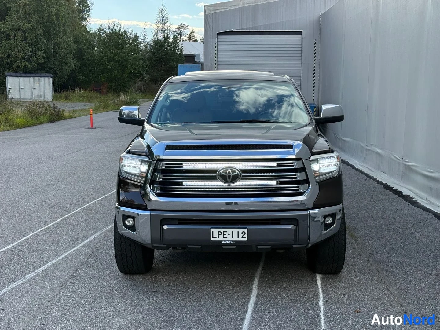 Edestä: Suora etunäkymä tummanruskeasta avolavasta: kromikehystetty etusäleikkö Toyota-merkillä, LED-päiväajovalot ja rekisteritunnus.
