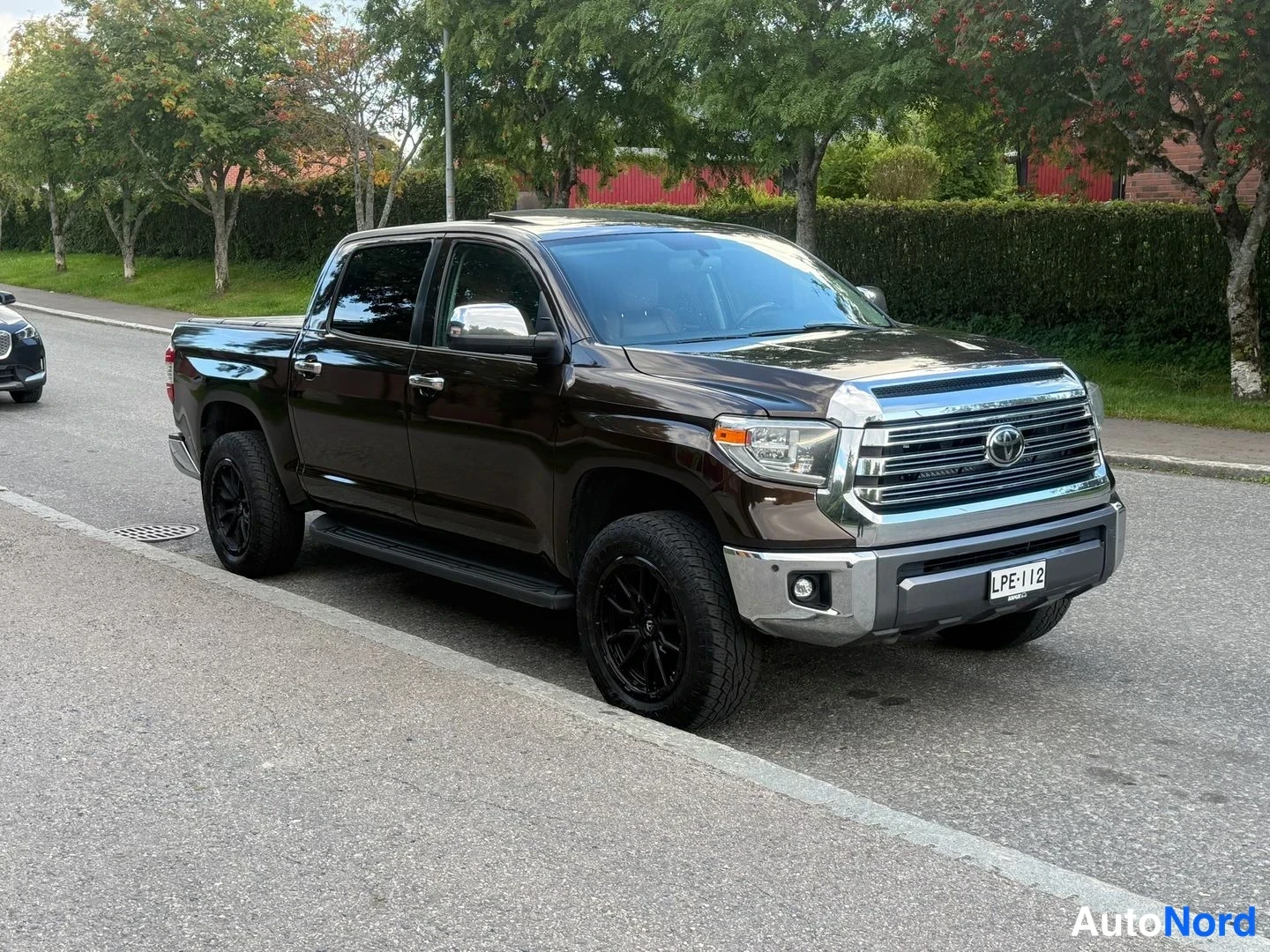 Etuviisto, nokka oikealle: Tummanruskea Toyota Tundra -nousija pysäköitynä kadunvarrella, kylki ja etuosa näkyvissä, mustat kevytmetallivanteet ja kromiin viimeistelty etusäleikkö.