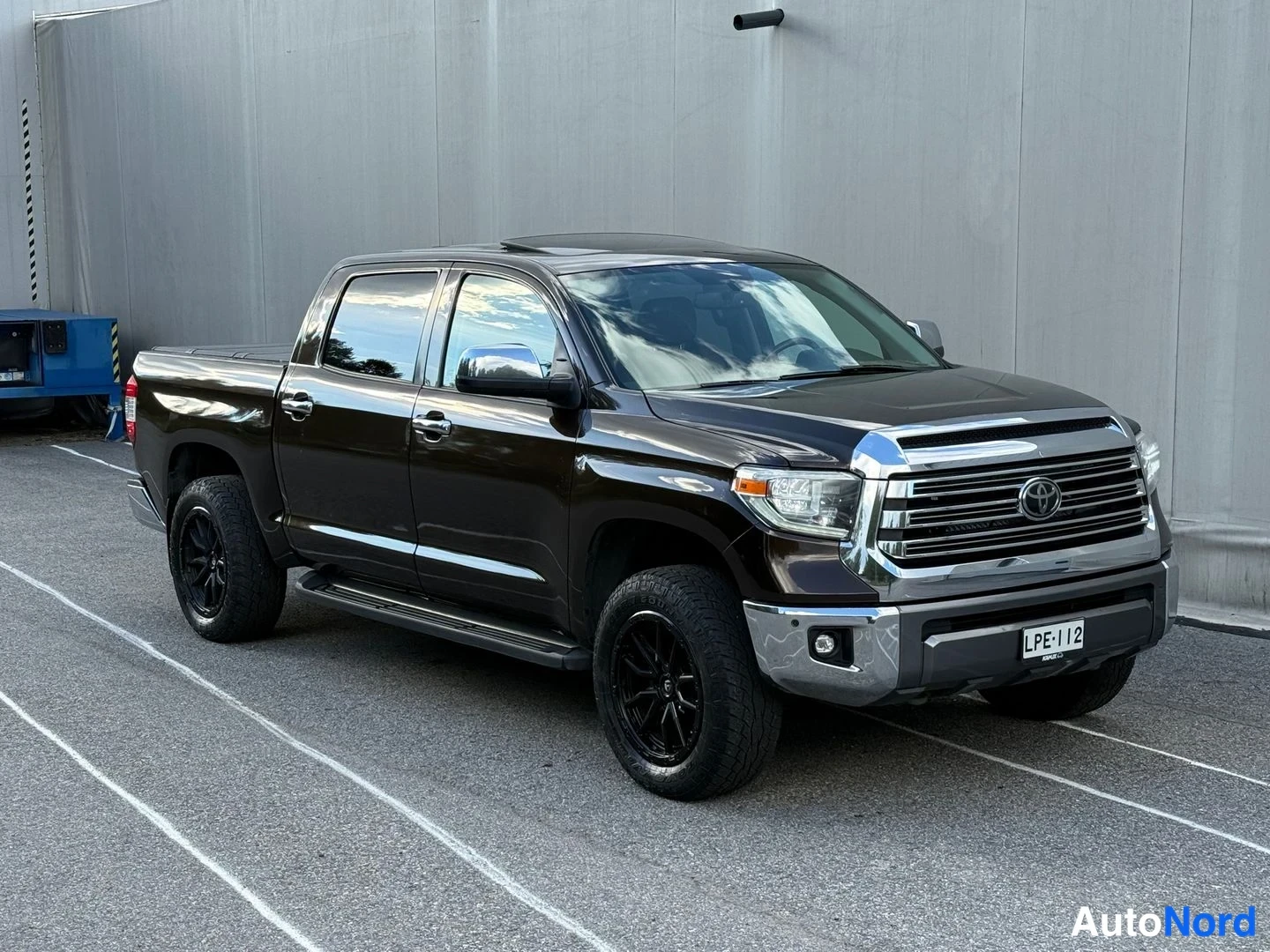Etuviisto, nokka oikealle: Toyota Tundra -nappikuorma-auto tummanruskeassa maalauksessa, etukolmasosa oikealta; kromikeula, mustat vanteet ja sivuastin näkyvissä.