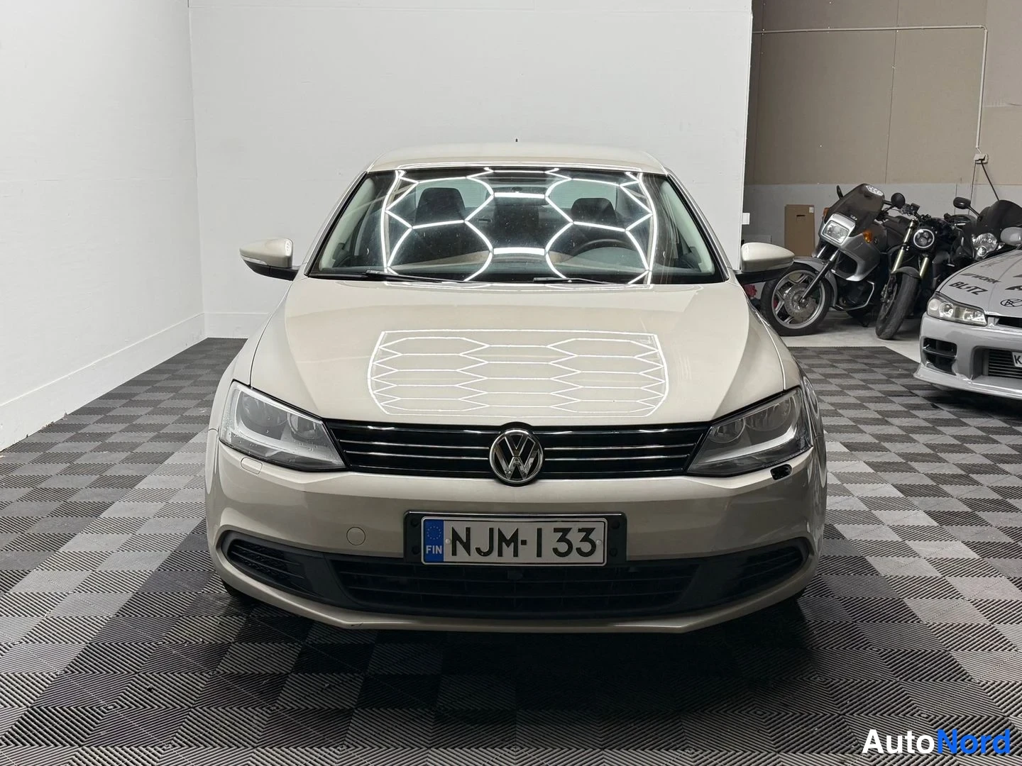 Edestä: Volkswagen Jetta -sedanin suora etänäkymä, vaaleanbeige konepelti, VW-tunnuksella varustettu etusäleikkö ja rekisterikilpi.