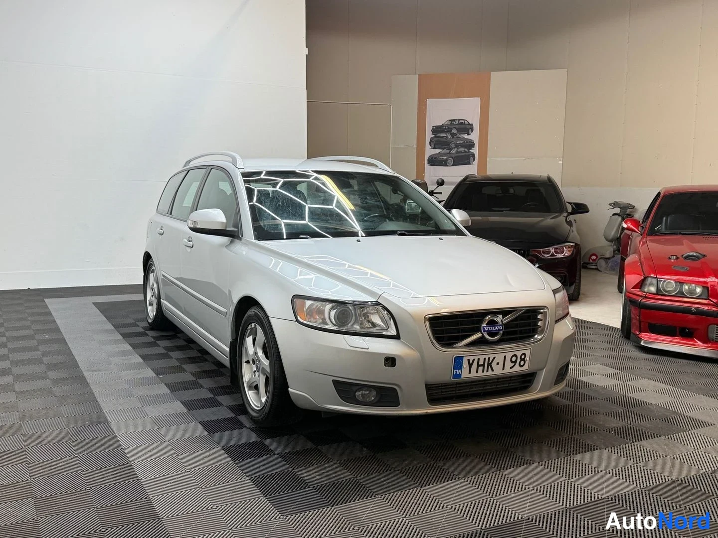 Etuviisto, nokka oikealle: Hopeinen Volvo V50 -farmari etuviistosta sisätilassa muiden autojen vieressä, rekisterikilpi.