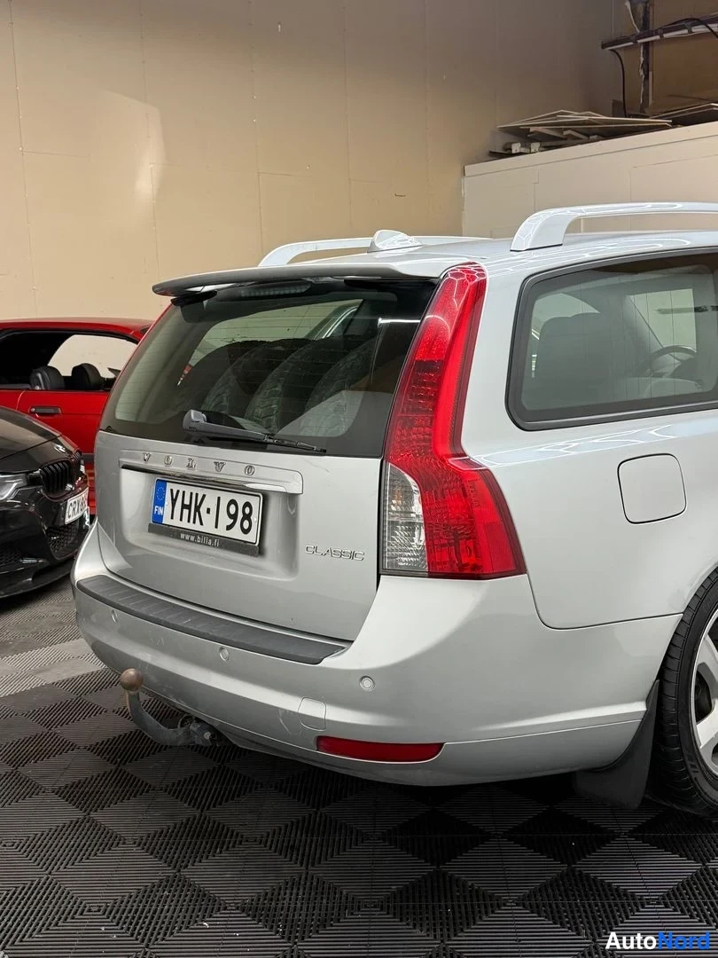 Takaviisto: Hopeanharmaa farmariauto takaa kuvattuna, rekisterikilpi, takavalot ja takavilkku näkyvät, konepellilla takana lähes kokonaan.