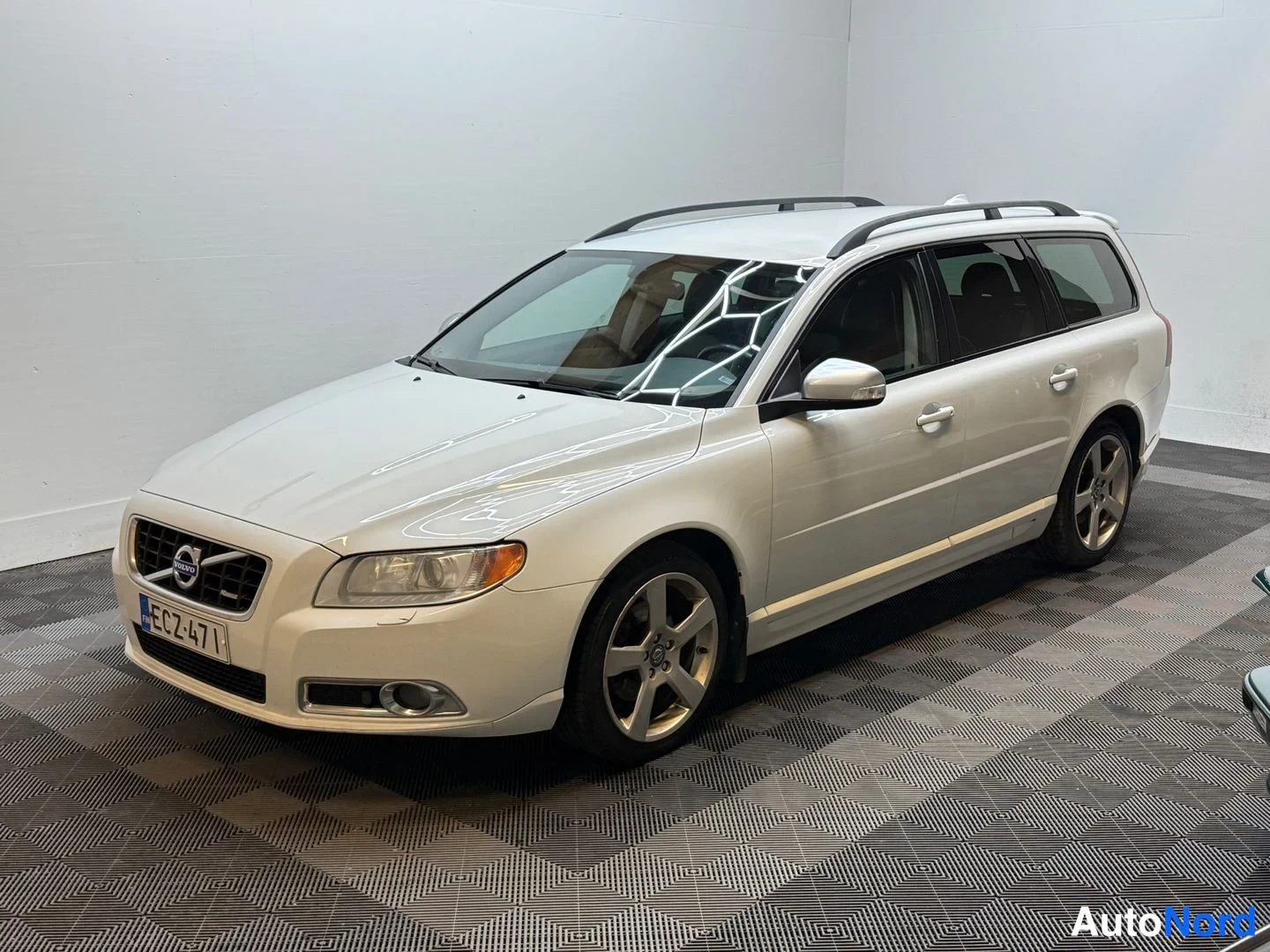 Etuviisto, nokka vasemmalle: Volvo V70 -farmari, valkoinen, etukulmasta kuvattuna sisätilassa. Rekisteritunnus, kattokaiteet ja viisikehäiset alumiinivanteet näkyvissä.