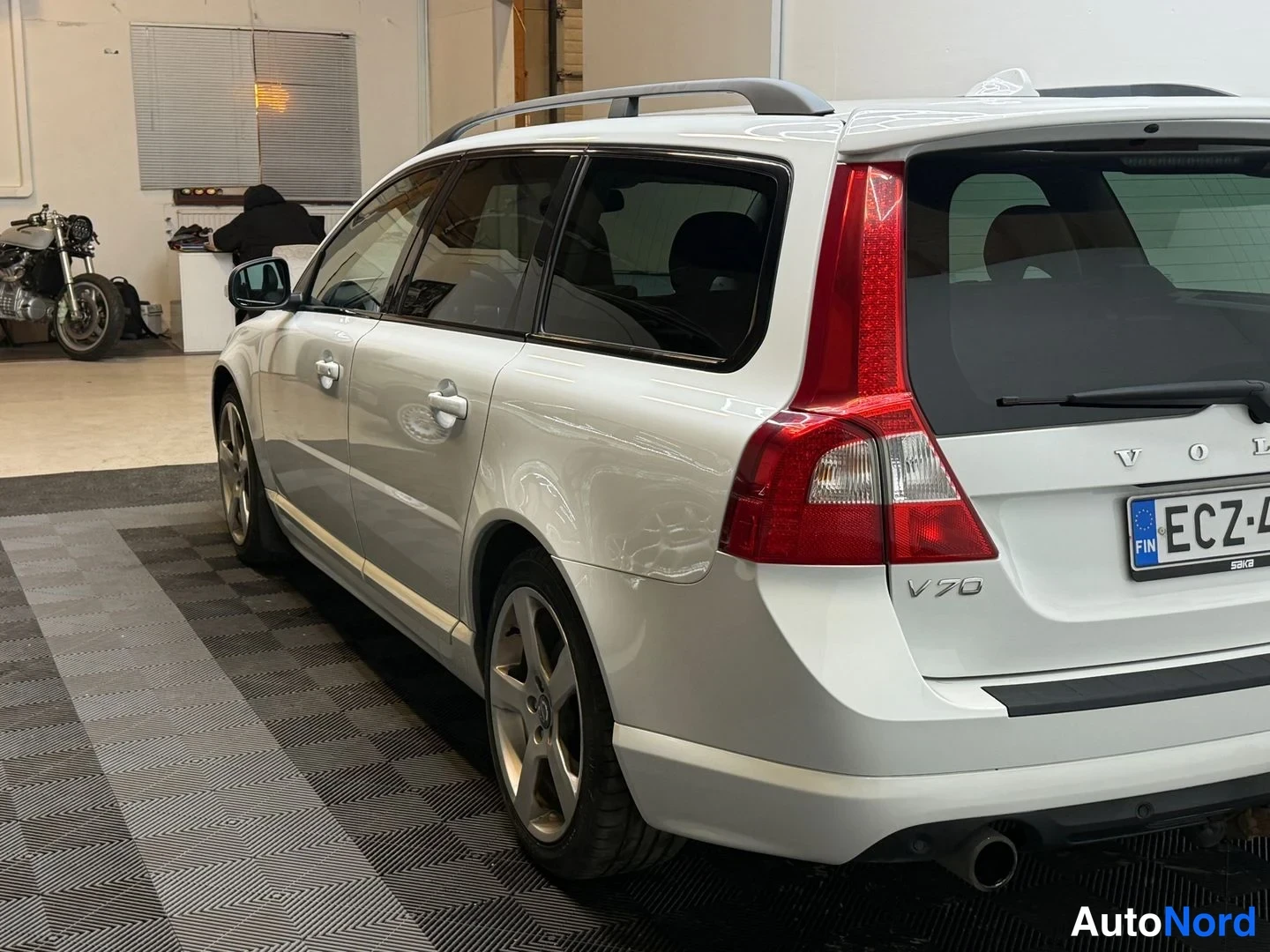 Takaviisto: Auton takakulma sisätilassa: valkoinen farmariauto, takavalot ja V70-merkintä näkyvät, rekisterikilpi.