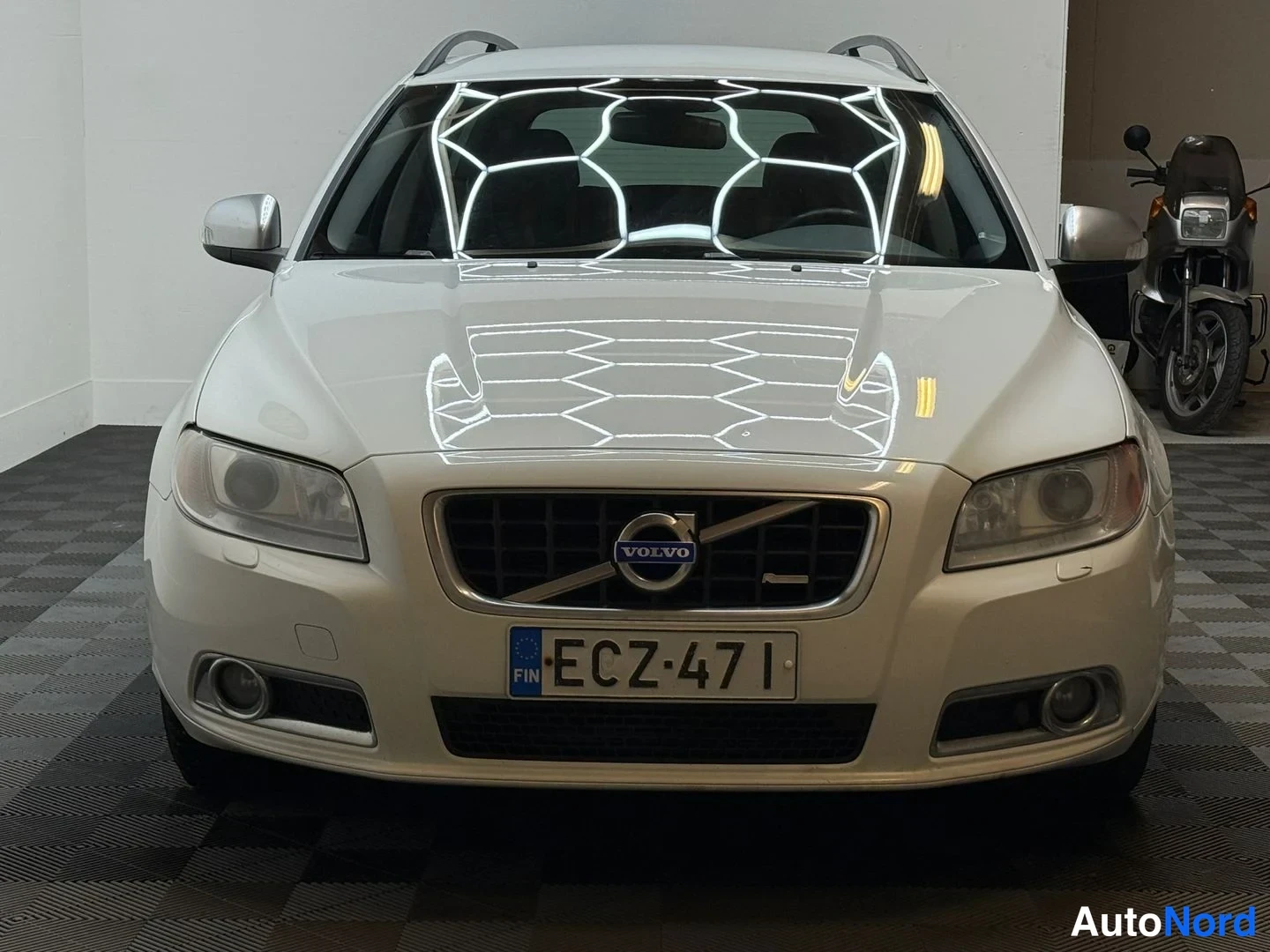 Edestä: Valkoisen Volvo V70 -farmarin suora etänäkymä, -rekisterikilpi ja kattokaiteet näkyvissä.
