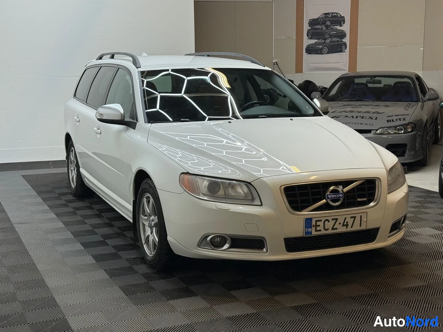 Etuviisto, nokka oikealle: Valkoinen Volvo V70 -farmari kolmeneljäsosan etunäkymä vasemmasta kyljestä, kattokaiteet ja -kilpi näkyvissä.
