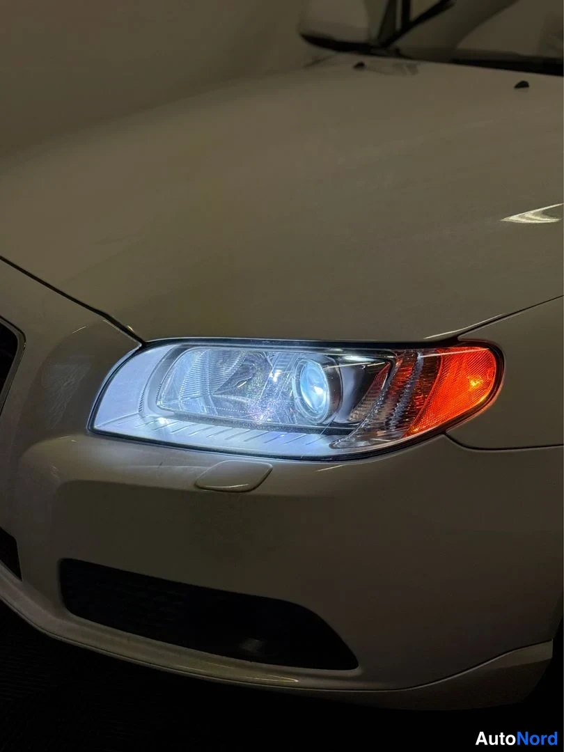 Edestä: Volvo V70:n etuosan lähikuva tummassa tilassa: valkoinen ajovalo ja oranssi suuntavilkku ovat päällä, etupuskurin ja konepellin reuna näkyvät.