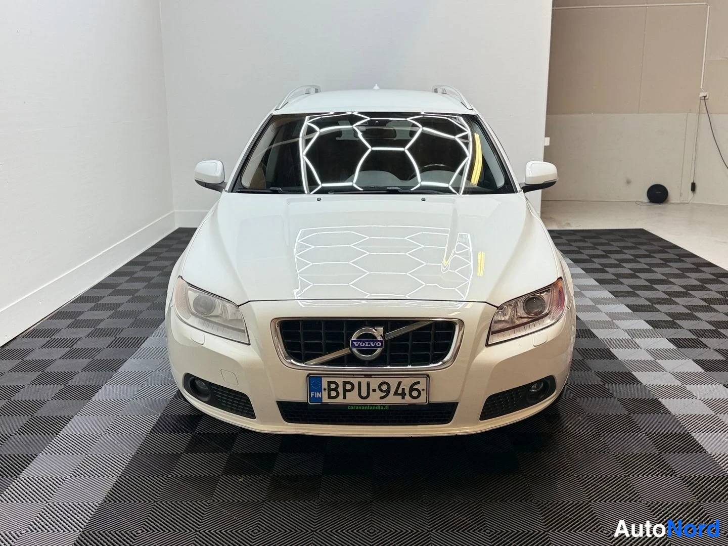 Edestä: Auton etupäätä kohti kuvattu valkoinen Volvo V70 sisätilassa, rekisterikilpi, kattokaiteet näkyvissä.